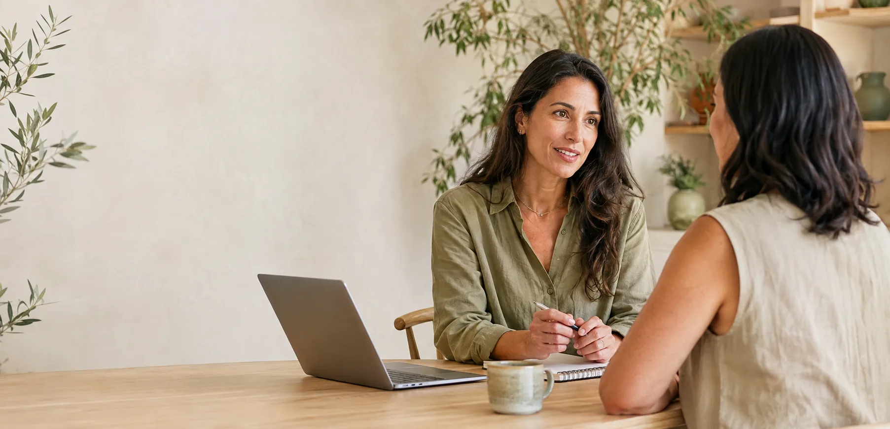 Coach en cliënt in gesprek aan een houten tafel met laptop en notitieboek
